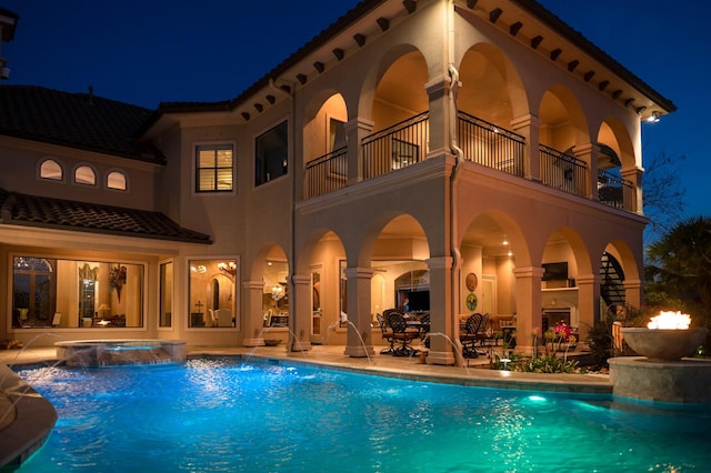 back of house at twilight with stucco siding, a patio, an outdoor pool, a balcony, and an in ground hot tub