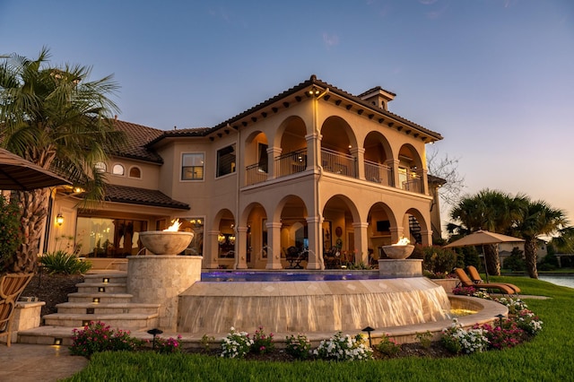 back of house at dusk with a balcony, a tiled roof, and stucco siding