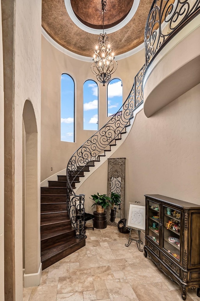 stairs with a tray ceiling, arched walkways, ornamental molding, stone finish floor, and a notable chandelier