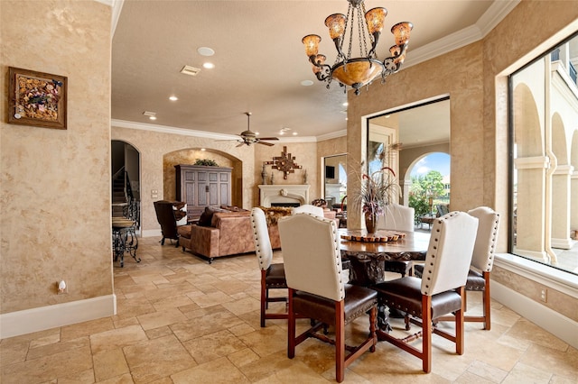 dining space featuring stone tile floors, baseboards, a lit fireplace, crown molding, and ceiling fan with notable chandelier