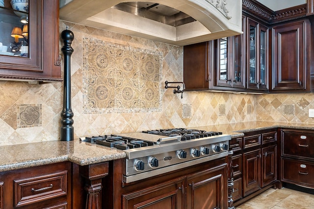 kitchen featuring backsplash, stone tile floors, stainless steel gas stovetop, glass insert cabinets, and light stone countertops