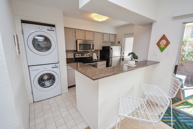 kitchen with tasteful backsplash, appliances with stainless steel finishes, stacked washer and clothes dryer, light tile patterned floors, and kitchen peninsula