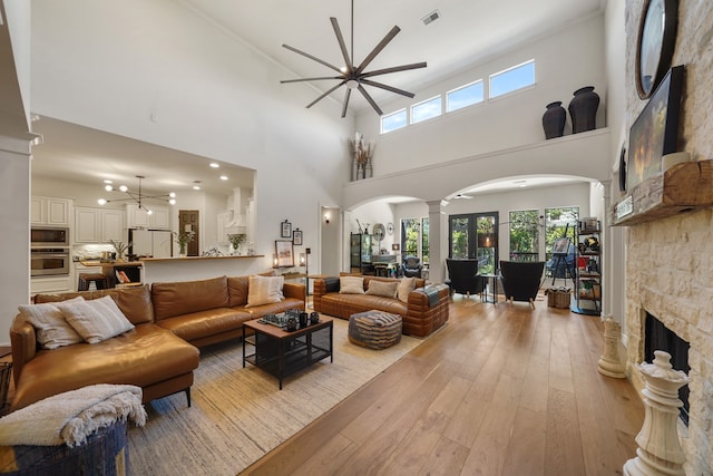 living room with decorative columns, light wood-type flooring, ceiling fan, a stone fireplace, and a towering ceiling