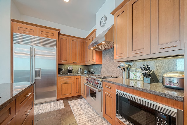 kitchen featuring dark stone countertops, tile patterned floors, tasteful backsplash, built in appliances, and wall chimney range hood