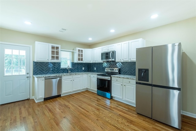 kitchen with light stone counters, appliances with stainless steel finishes, glass insert cabinets, white cabinetry, and light wood-type flooring