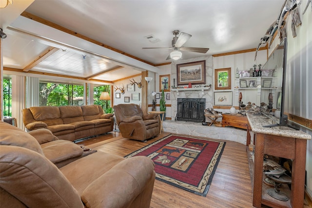 living room with crown molding, beamed ceiling, light wood-type flooring, ceiling fan, and a stone fireplace