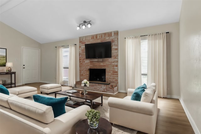 living room featuring light wood-type flooring, brick wall, vaulted ceiling, rail lighting, and a fireplace