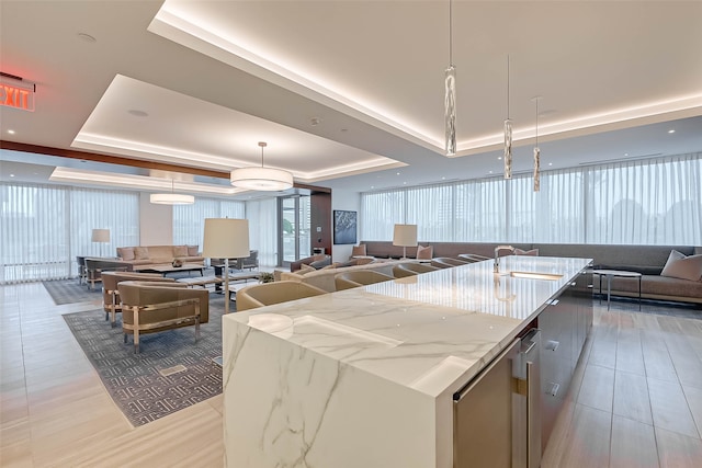 kitchen with a raised ceiling, hanging light fixtures, an island with sink, light stone counters, and sink