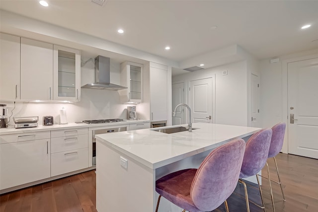 kitchen with sink, dark hardwood / wood-style flooring, a kitchen island with sink, wall chimney range hood, and white cabinets