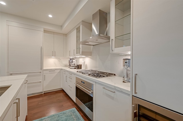 kitchen with appliances with stainless steel finishes, dark wood-type flooring, light stone counters, white cabinetry, and wall chimney range hood