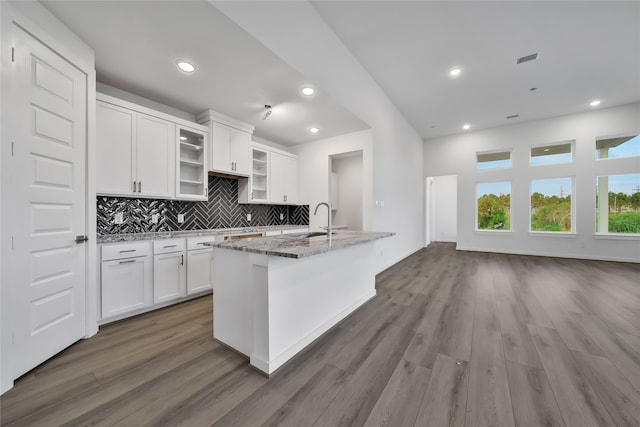 kitchen with a center island with sink, decorative backsplash, hardwood / wood-style flooring, light stone countertops, and white cabinetry