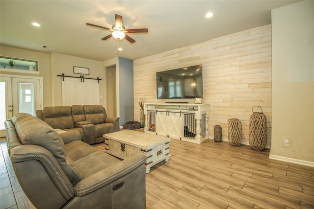 living room featuring ceiling fan, french doors, wooden walls, light wood-type flooring, and a barn door