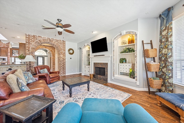 living room featuring ceiling fan, built in features, a tiled fireplace, hardwood / wood-style flooring, and a textured ceiling