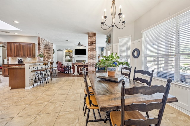 tiled dining room with ceiling fan with notable chandelier, brick wall, and a brick fireplace