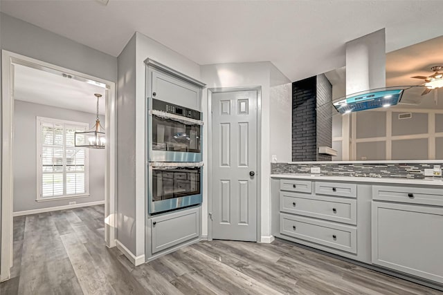 kitchen featuring ceiling fan with notable chandelier, island exhaust hood, light wood finished floors, and stainless steel double oven