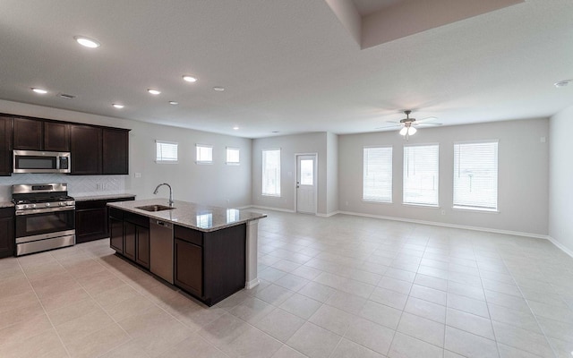 kitchen with sink, dark brown cabinets, a center island with sink, stainless steel appliances, and light stone countertops