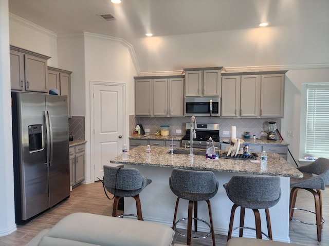 kitchen featuring appliances with stainless steel finishes, backsplash, gray cabinetry, sink, and an island with sink