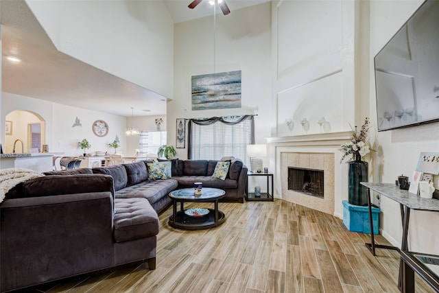 living room with a towering ceiling, a tiled fireplace, light hardwood / wood-style flooring, and ceiling fan with notable chandelier