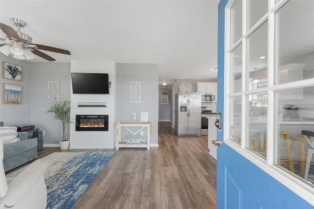 living room featuring wood finished floors, a glass covered fireplace, a ceiling fan, and baseboards