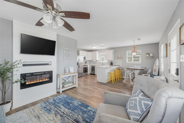 living area featuring light wood finished floors, visible vents, a ceiling fan, and a glass covered fireplace