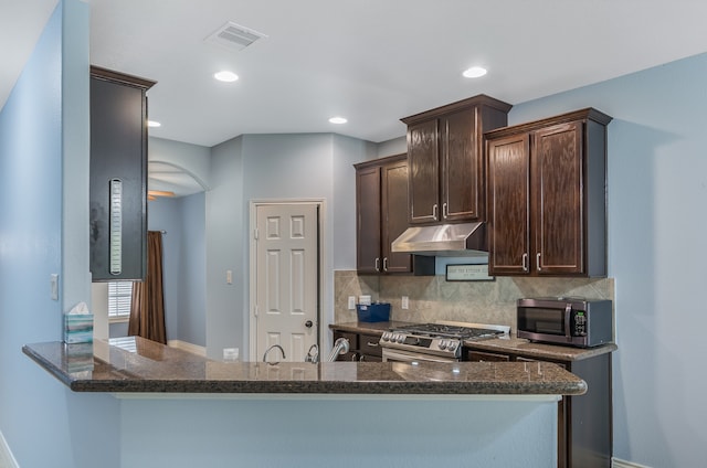 kitchen with appliances with stainless steel finishes, kitchen peninsula, dark stone countertops, and dark brown cabinets