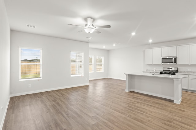 kitchen featuring ceiling fan, light hardwood / wood-style floors, appliances with stainless steel finishes, white cabinets, and an island with sink