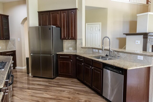 kitchen featuring stainless steel appliances, a peninsula, a sink, backsplash, and light stone countertops