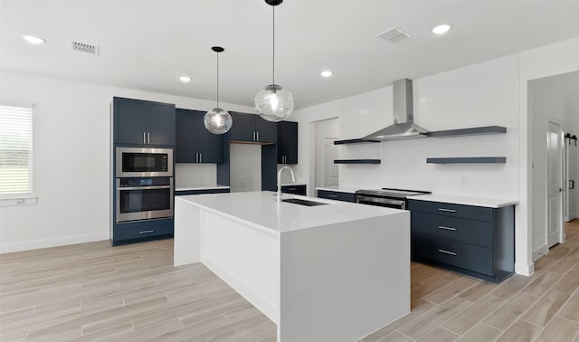 kitchen with sink, hanging light fixtures, a center island with sink, stainless steel appliances, and wall chimney range hood