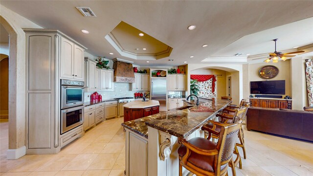 kitchen featuring light tile patterned flooring, custom range hood, appliances with stainless steel finishes, a raised ceiling, and a center island with sink