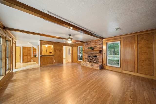 unfurnished living room featuring a textured ceiling, beamed ceiling, wooden walls, a brick fireplace, and light wood-type flooring