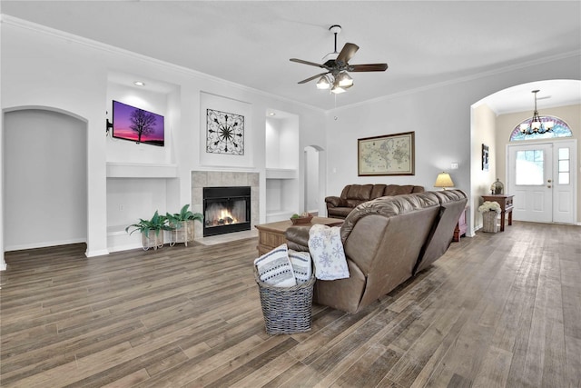 living room featuring built in shelves, crown molding, a tiled fireplace, dark wood-type flooring, and ceiling fan