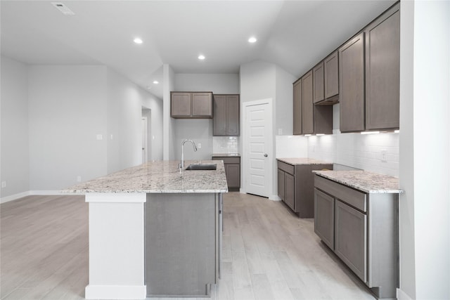 kitchen featuring tasteful backsplash, an island with sink, light stone counters, light wood-style floors, and a sink