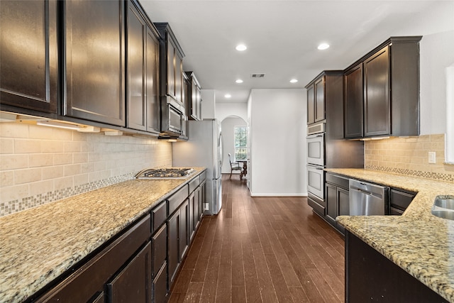 kitchen with light stone counters, stainless steel appliances, dark brown cabinets, dark wood-type flooring, and decorative backsplash