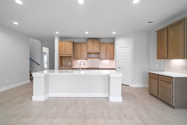 kitchen with light wood-type flooring, sink, backsplash, and an island with sink