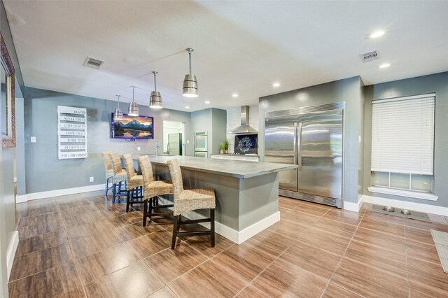 kitchen with built in fridge, a breakfast bar area, wall chimney exhaust hood, pendant lighting, and a spacious island