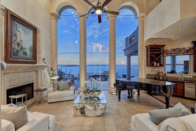 tiled living room featuring a fireplace, a water view, a high ceiling, ceiling fan, and ornate columns