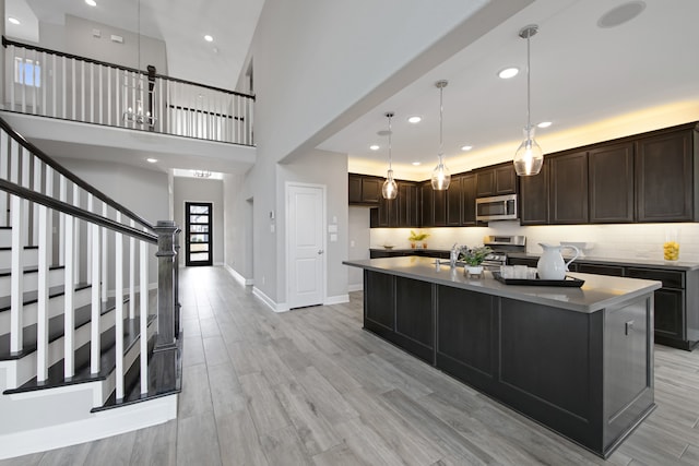 kitchen featuring light wood-type flooring, a towering ceiling, an island with sink, dark brown cabinets, and stainless steel appliances