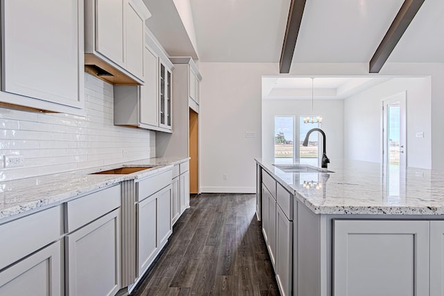 kitchen featuring sink, a kitchen island with sink, dark hardwood / wood-style flooring, light stone counters, and beamed ceiling