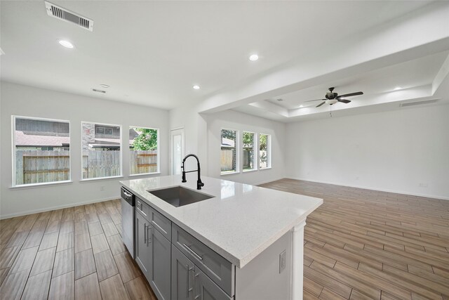 kitchen with light wood-type flooring, sink, ceiling fan, a raised ceiling, and a center island with sink