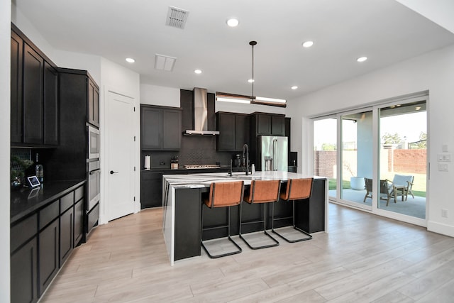 kitchen with backsplash, decorative light fixtures, stainless steel appliances, a center island with sink, and wall chimney range hood