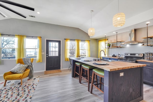 kitchen with stainless steel electric stove, wall chimney exhaust hood, a kitchen island with sink, light hardwood / wood-style floors, and wooden counters