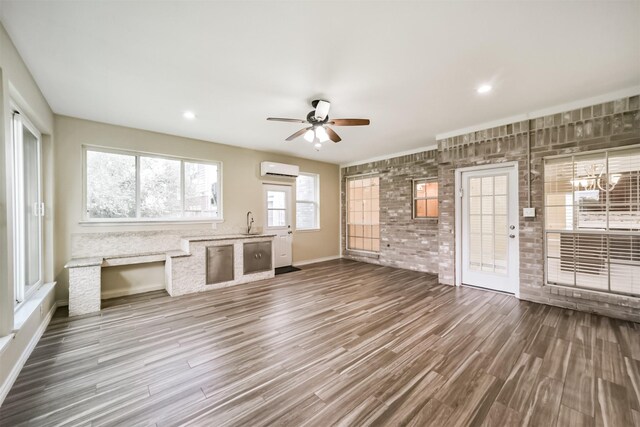 unfurnished living room with ceiling fan with notable chandelier, a sink, dark wood finished floors, a wall unit AC, and brick wall