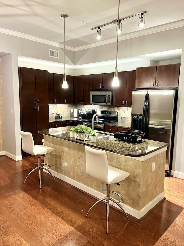 kitchen with appliances with stainless steel finishes, hardwood / wood-style flooring, dark stone counters, and a kitchen island with sink