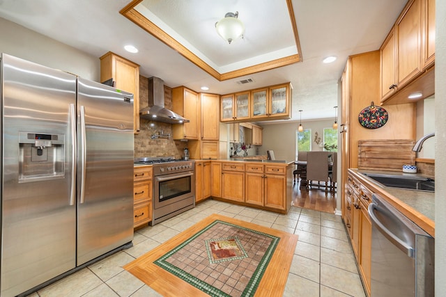 kitchen with a raised ceiling, appliances with stainless steel finishes, a sink, wall chimney range hood, and a peninsula