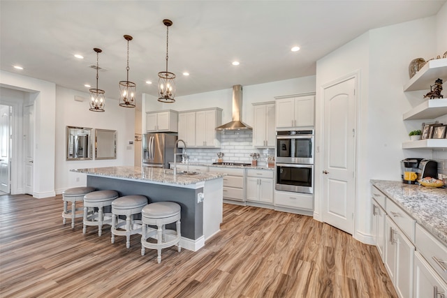 kitchen with a kitchen island with sink, white cabinets, wall chimney range hood, light hardwood / wood-style flooring, and appliances with stainless steel finishes