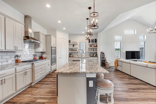 kitchen featuring an island with sink, wall chimney exhaust hood, hanging light fixtures, and sink