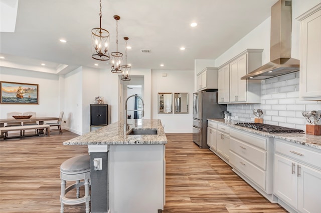 kitchen featuring a chandelier, light hardwood / wood-style floors, sink, wall chimney range hood, and a center island with sink