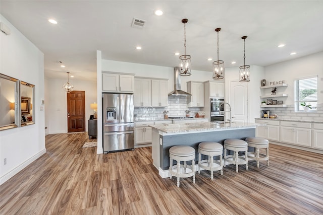 kitchen featuring pendant lighting, a kitchen island with sink, wall chimney exhaust hood, stainless steel appliances, and light wood-type flooring