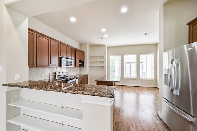 kitchen featuring light hardwood / wood-style flooring, backsplash, appliances with stainless steel finishes, kitchen peninsula, and dark stone counters