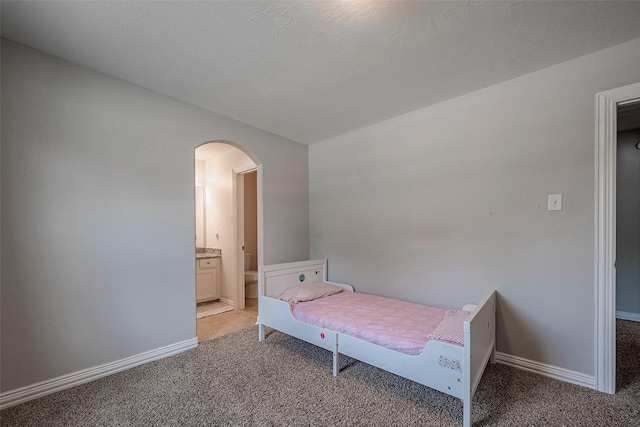 bedroom featuring baseboards, arched walkways, ensuite bathroom, and light carpet